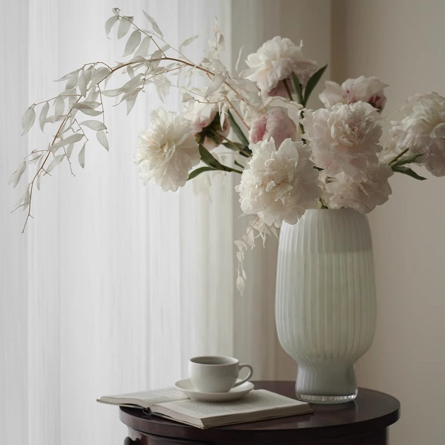 White ribbed ceramic vase with soft pink peonies on a wooden table by a sunlit window – minimalist spring décor style