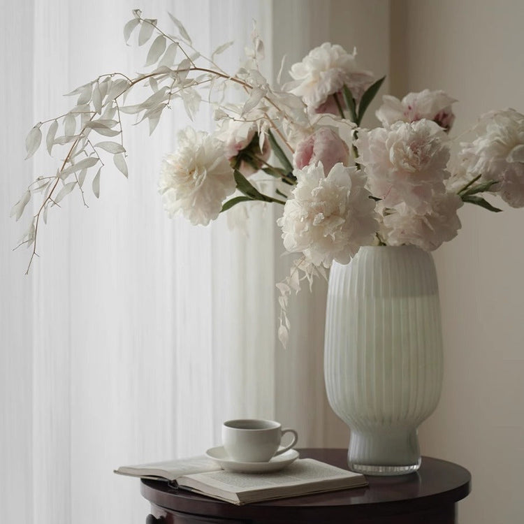 White ribbed ceramic vase with soft pink peonies on a wooden table by a sunlit window – minimalist spring décor style